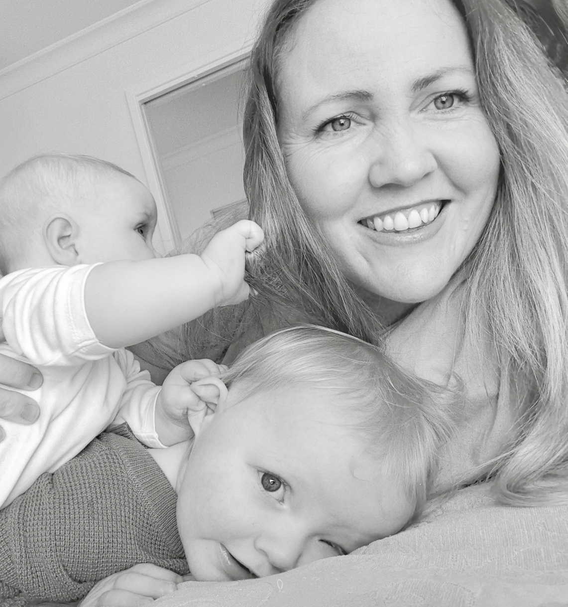 A woman smiling while holding two babies, one of whom is pulling her hair, and the other is laying beside her, both looking at the camera.